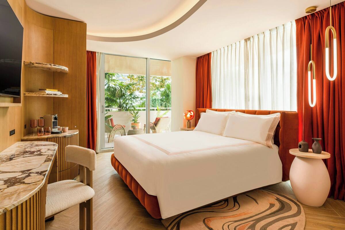 A modern hotel room with a bed, a desk, a chair, a TV, a balcony with plants, and a view of greenery.