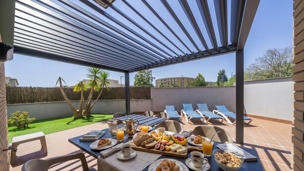 Eine Terrasse im Freien mit einem Frühstückstisch, der mit Croissants, Obst und Getränken gedeckt ist, unter einer Pergola mit Blick auf den Garten und Gebäude im Hintergrund.