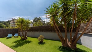 Ein gepflegter Garten mit Kunstgrashalm-Bepflanzung, zwei blauen Liegestühlen und Palmen.