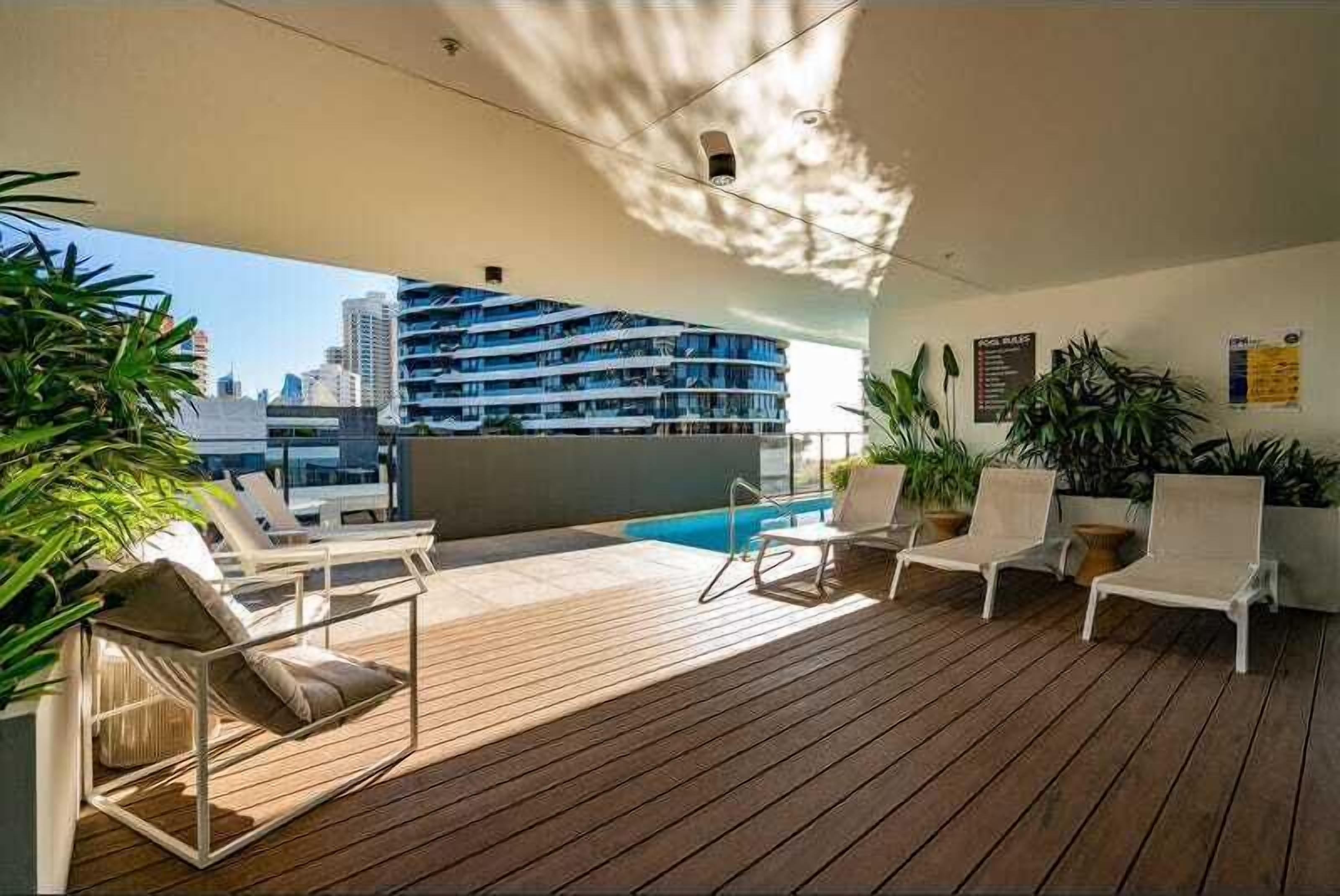 A sunlit outdoor deck with lounge chairs and a pool, overlooking a cityscape.