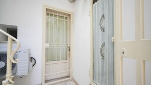 Un pasillo estrecho con una puerta blanca que tiene una reja metálica, una ventana con cortinas decorativas y un equipo de aire acondicionado montado en la pared.