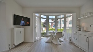 Eine moderne Küche mit weißen Schränken und einem eingebauten Backofen, ein Essbereich mit rundem Tisch und weißen Stühlen sowie ein an der Wand befestigter Fernseher.