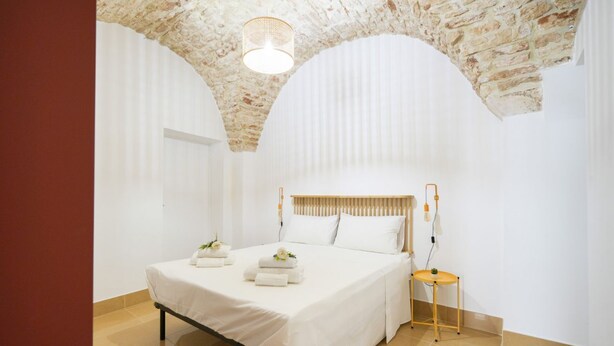 Een slaapkamer met een bed, witte lakens, een houten hoofdbord, een nachtkastje en een hanglamp.
