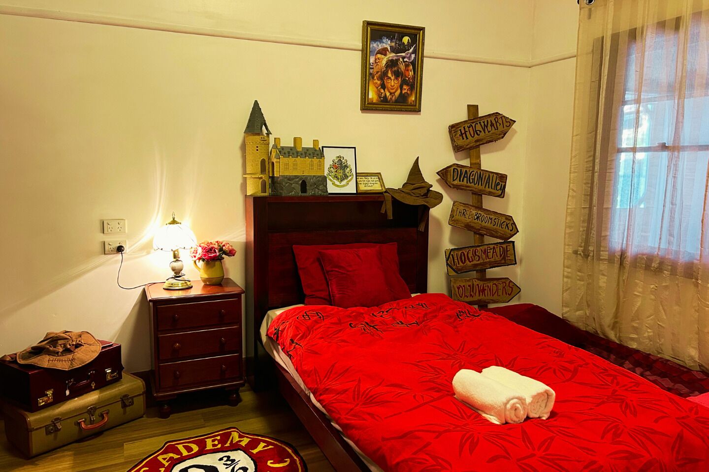 Een slaapkamer met een rode sprei, een houten hoofdbord, een nachtkastje met een lamp en bloemen, een koffer, en een bord met de routebeschrijvingen voor Hogwarts.