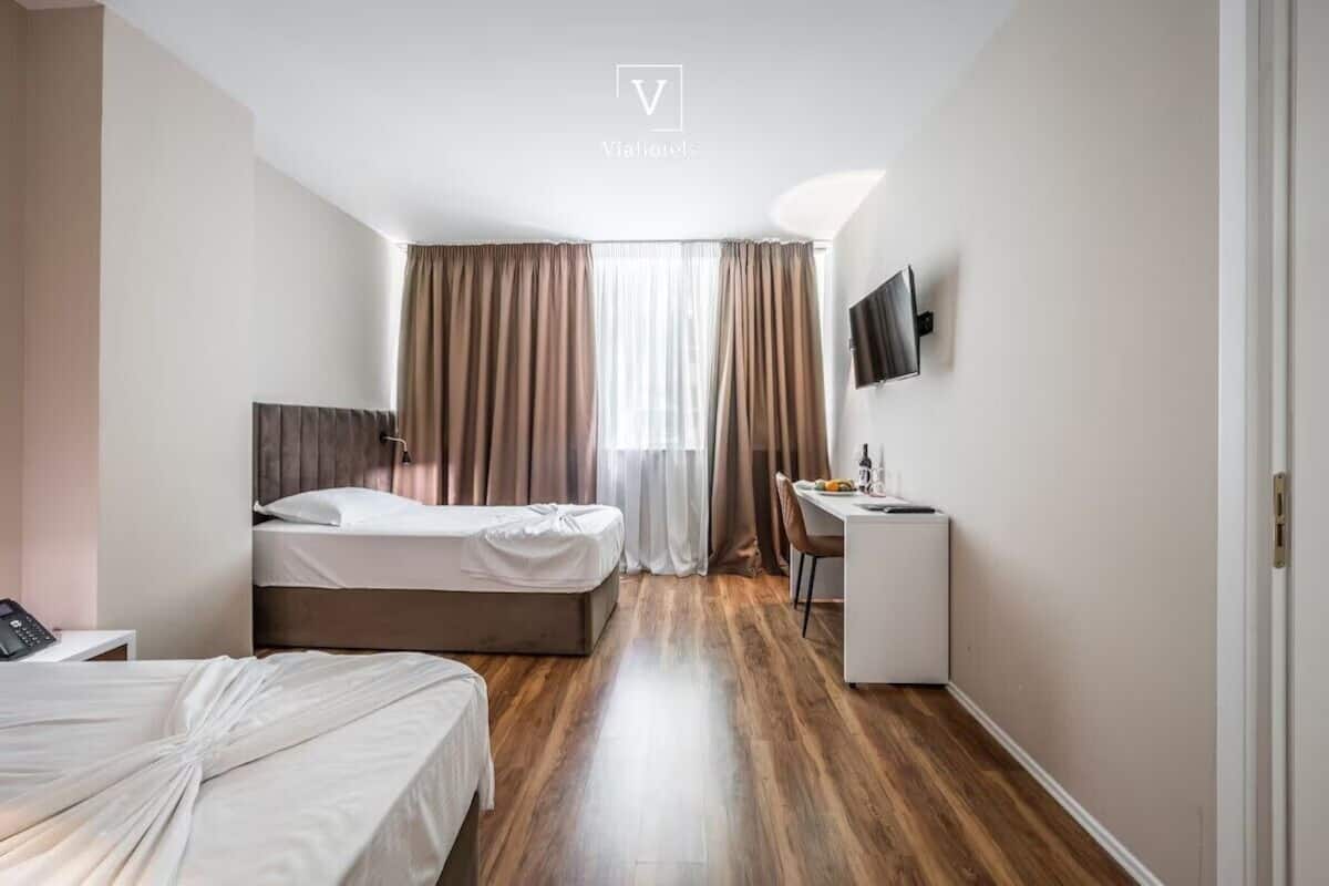 Habitación de hotel con cama, escritorio, silla, televisor montado en la pared y ventana con cortinas.