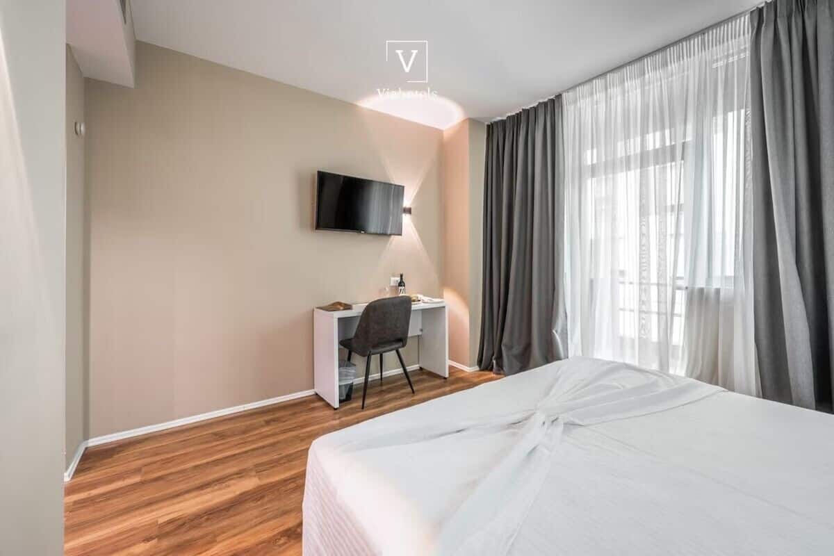 Habitación de hotel con cama, escritorio, silla, televisor montado en la pared y ventana con cortinas.