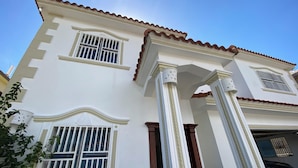 Un edificio de dos plantas con paredes blancas y techo de tejas rojas. Cuenta con ventanas grandes con barrotes blancos, columnas decorativas y un garaje con un coche estacionado.