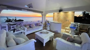 Un espacio lounge moderno al aire libre con mobiliario blanco, una barra y vistas al océano al atardecer.