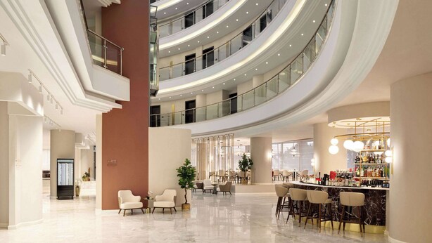 Un vestíbulo de hotel de varios niveles con una zona de bar central, arreglos de asientos y una escalera moderna.
