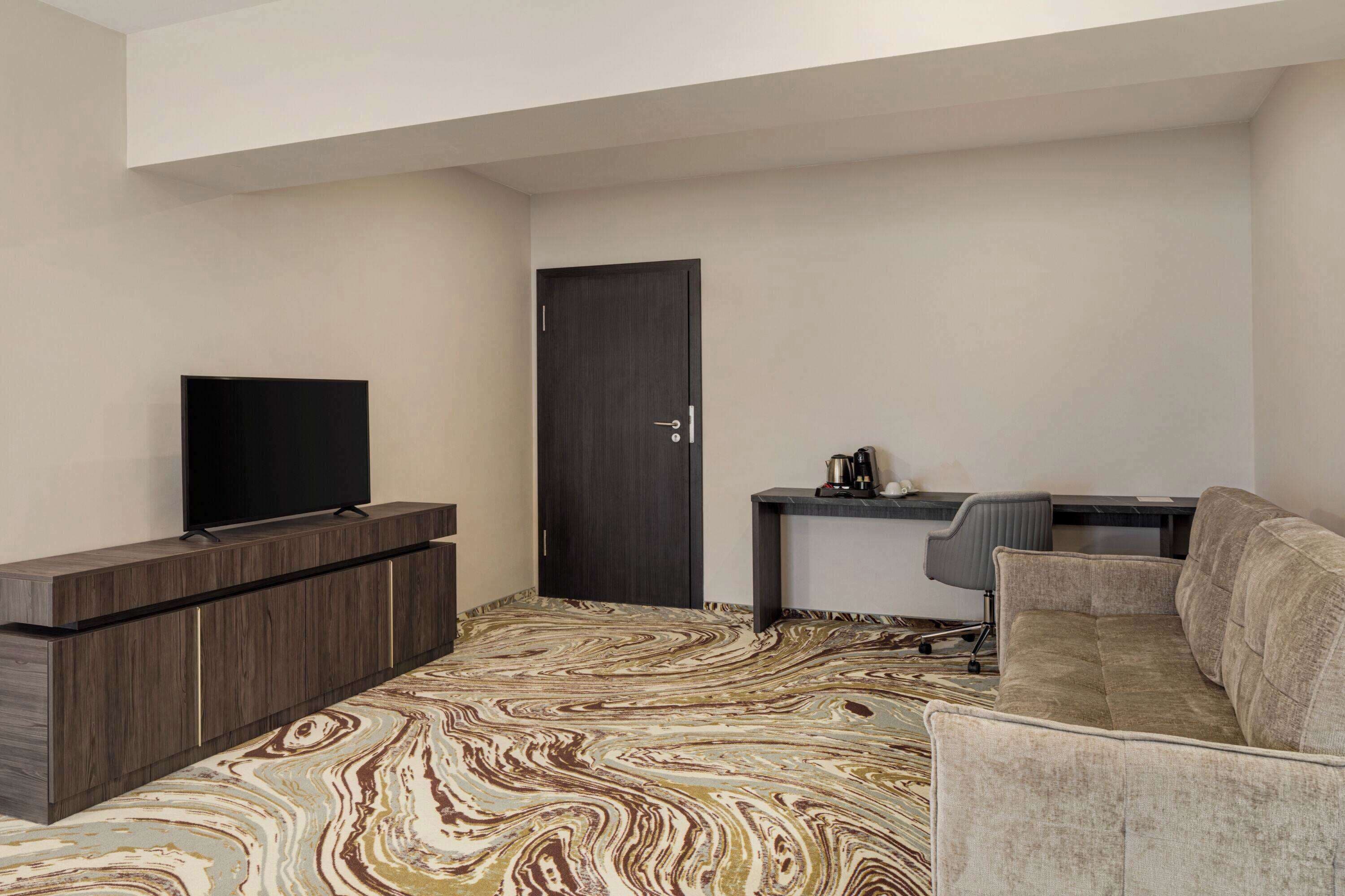 Ein modernes Zimmer mit Flachbildfernseher auf einer Holzkassette, einer dunklen Tür, einem kleinen Schreibtisch mit Stuhl und einem beigefarbenen Sofa.