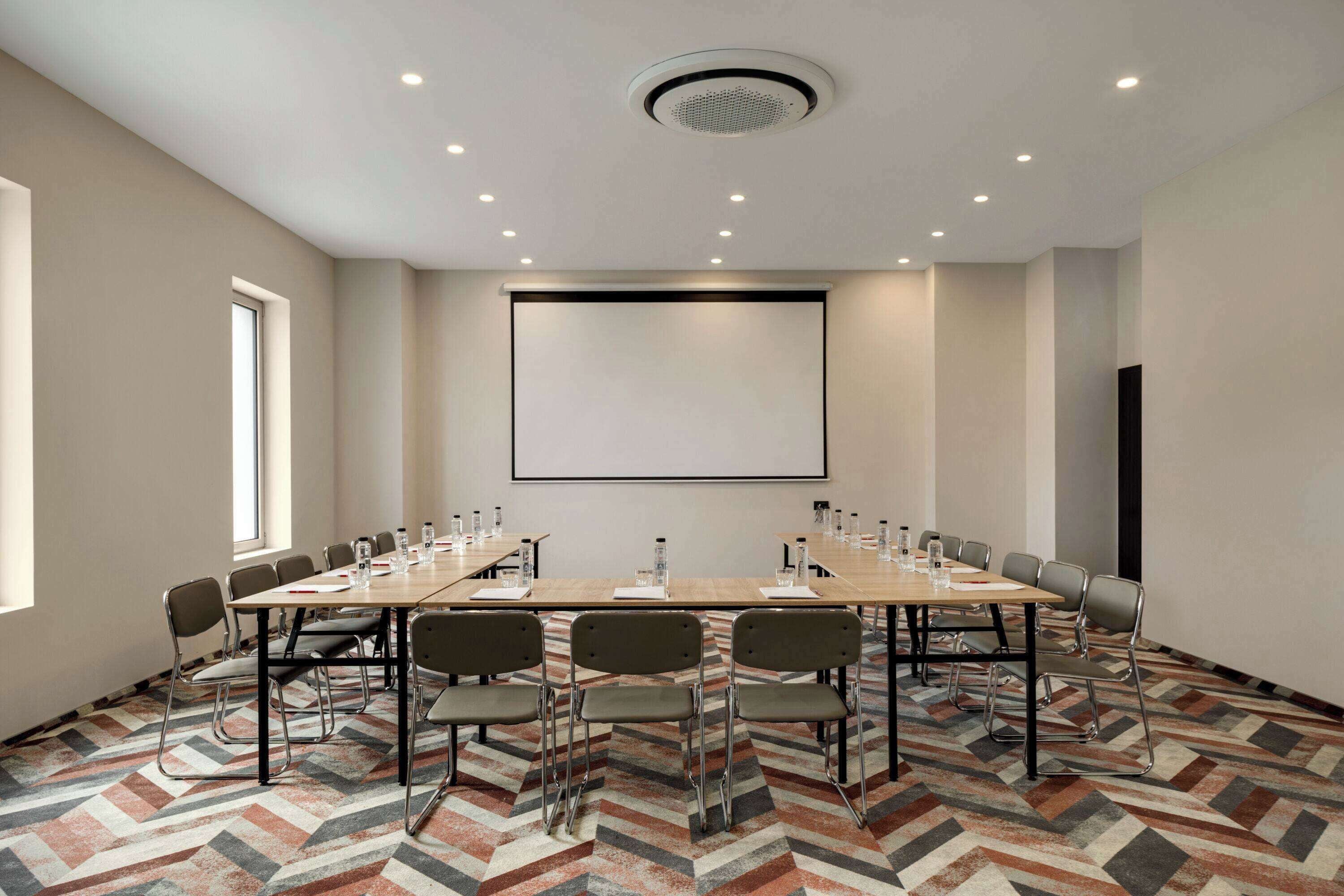 Un salón de conferencias con una mesa grande, sillas, una pantalla de proyector y una alfombra estampada.