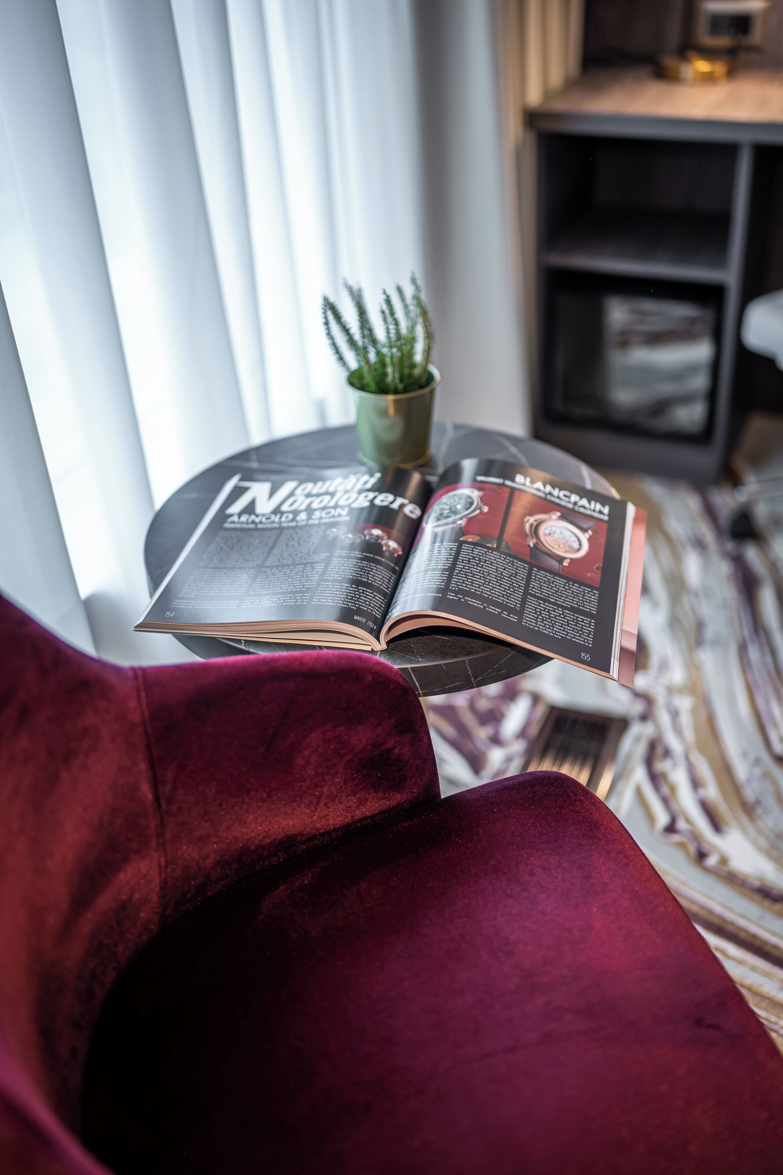 Una revista abierta sobre una mesa pequeña, una planta en maceta y una silla roja en una habitación con una ventana y un estante.