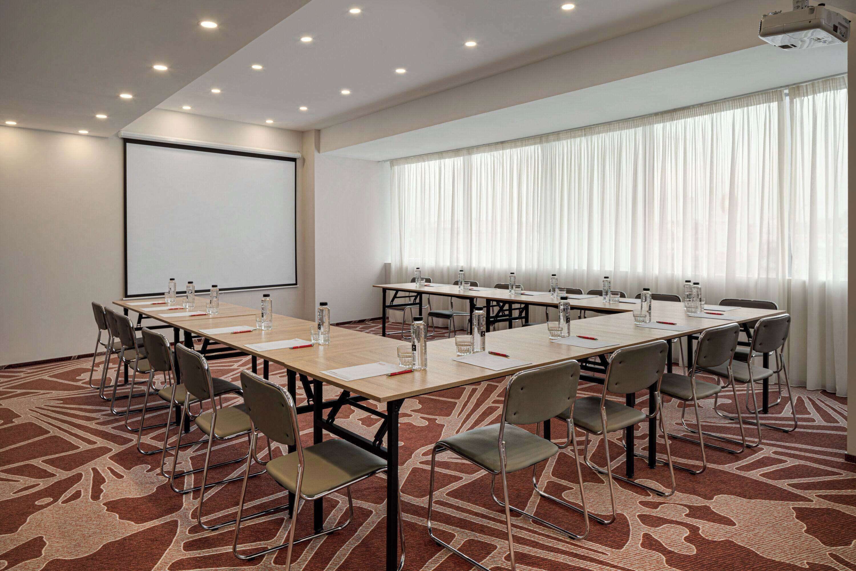 Une salle de conférence équipée d’une grande table, de chaises, d’un écran de projection et d’une grande fenêtre avec des rideaux.