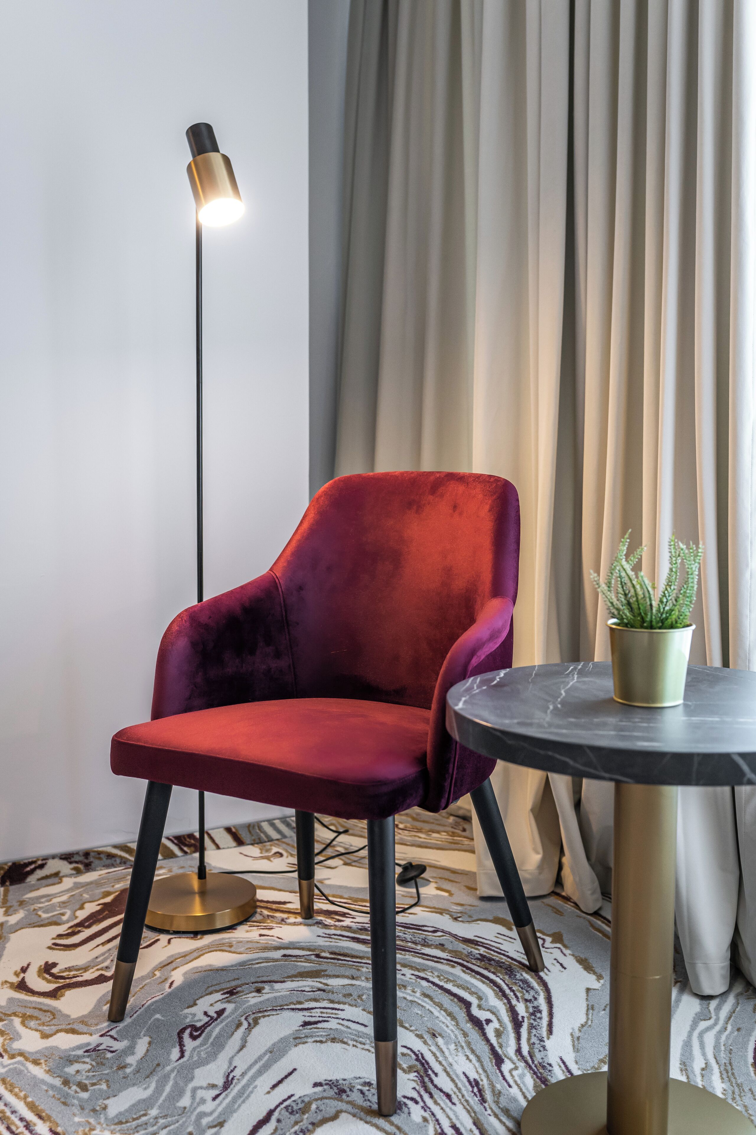Ein roter Samtstuhl, eine Stehlampe, ein kleiner Tisch mit einem Blumentopf und ein gemusterter Teppich in einem Raum mit Vorhang und Wand.