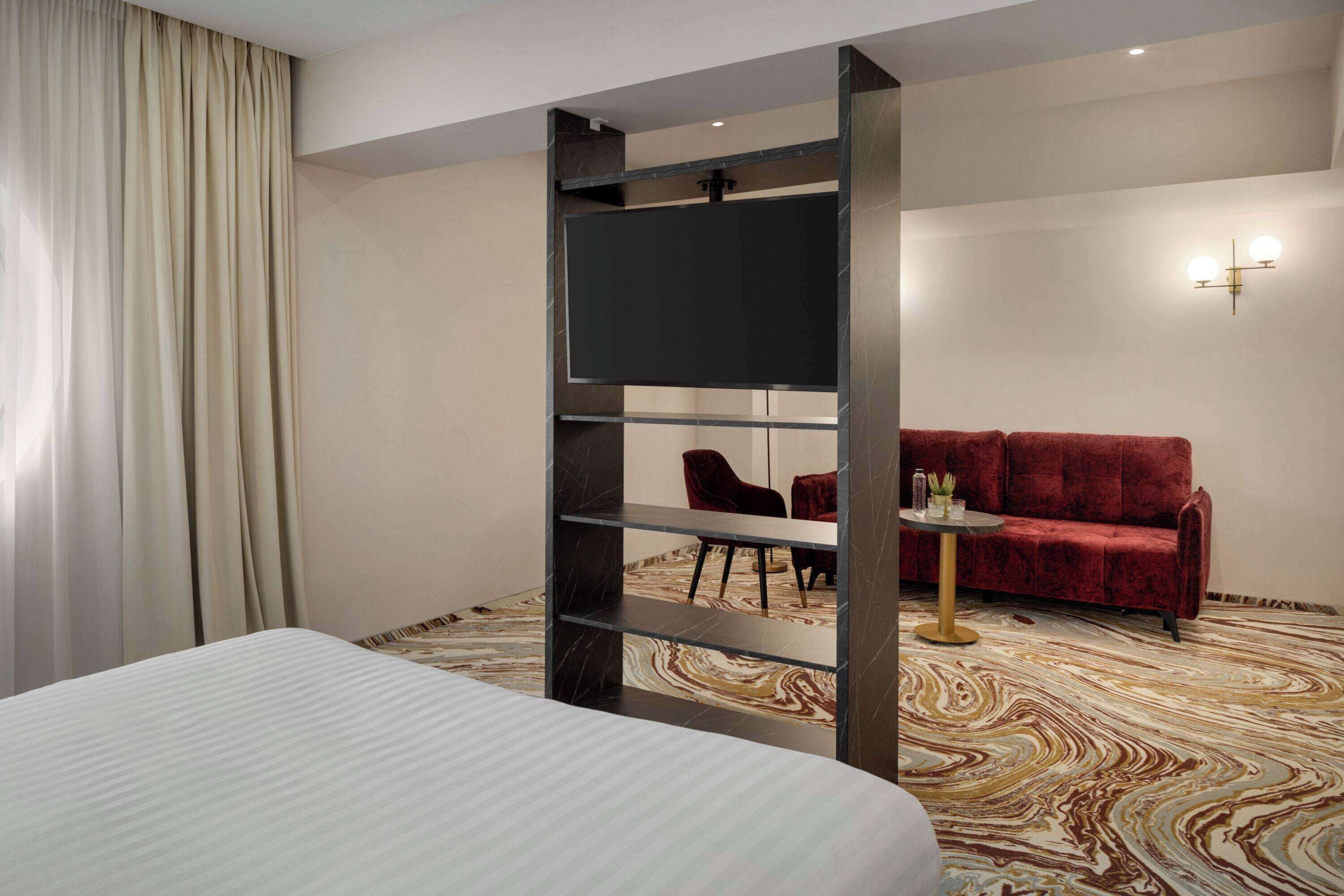 Een hotelkamer met een bed, een aan de muur bevestigde televisie, een rode bank, een tafeltje en een kamerbreed tapijt met een patroon.