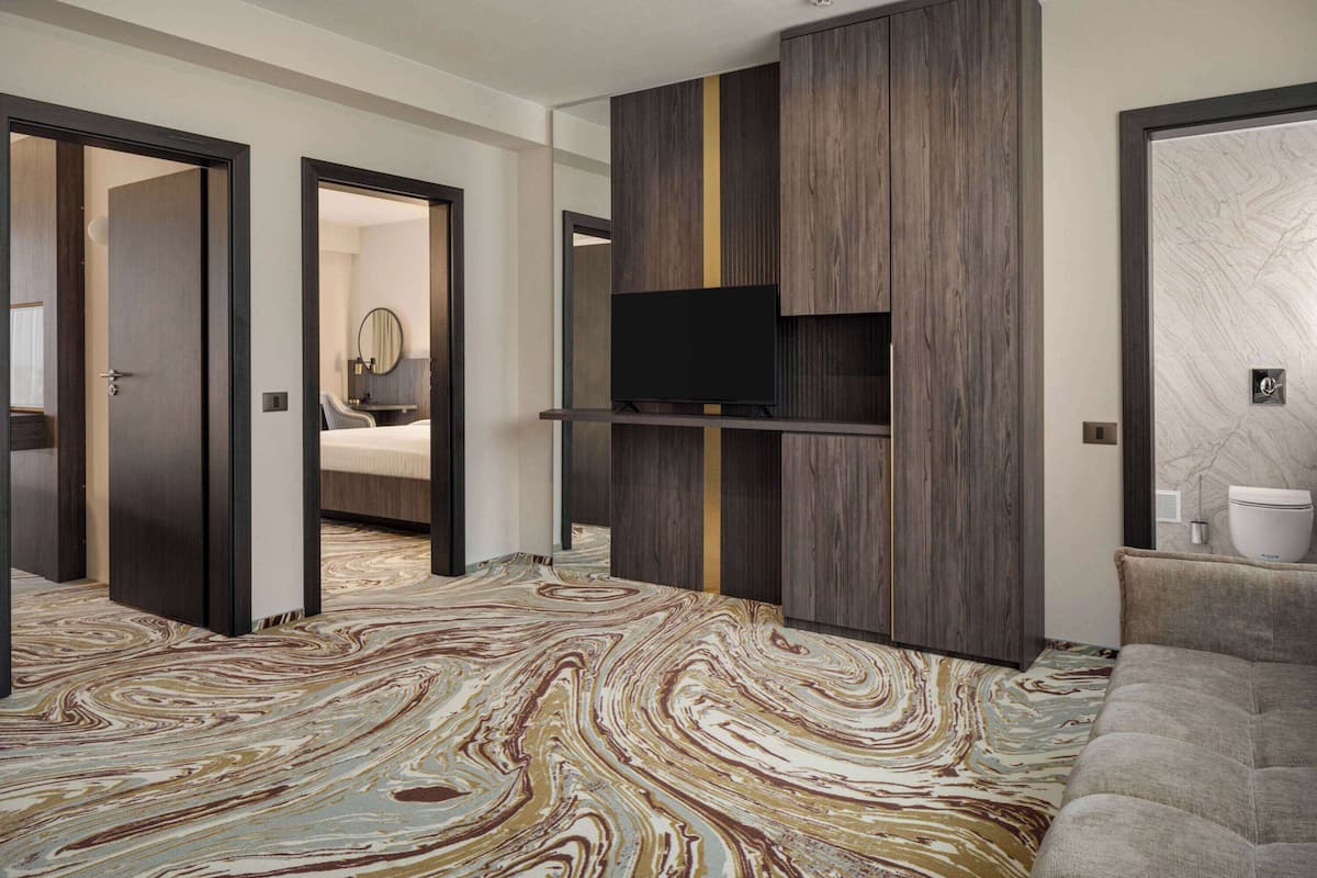 Una habitación de hotel moderna con piso de diseño de mármol, un televisor de pantalla plana montado en un panel de madera, un sofá y un baño con pared de mármol.