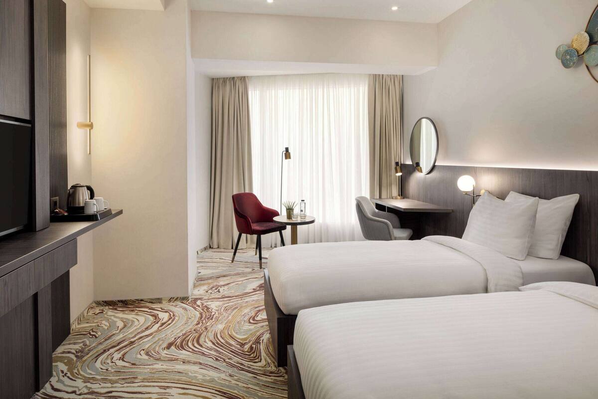 Habitación de hotel con dos camas, un escritorio, una silla, un televisor y un ventanal grande con cortinas.