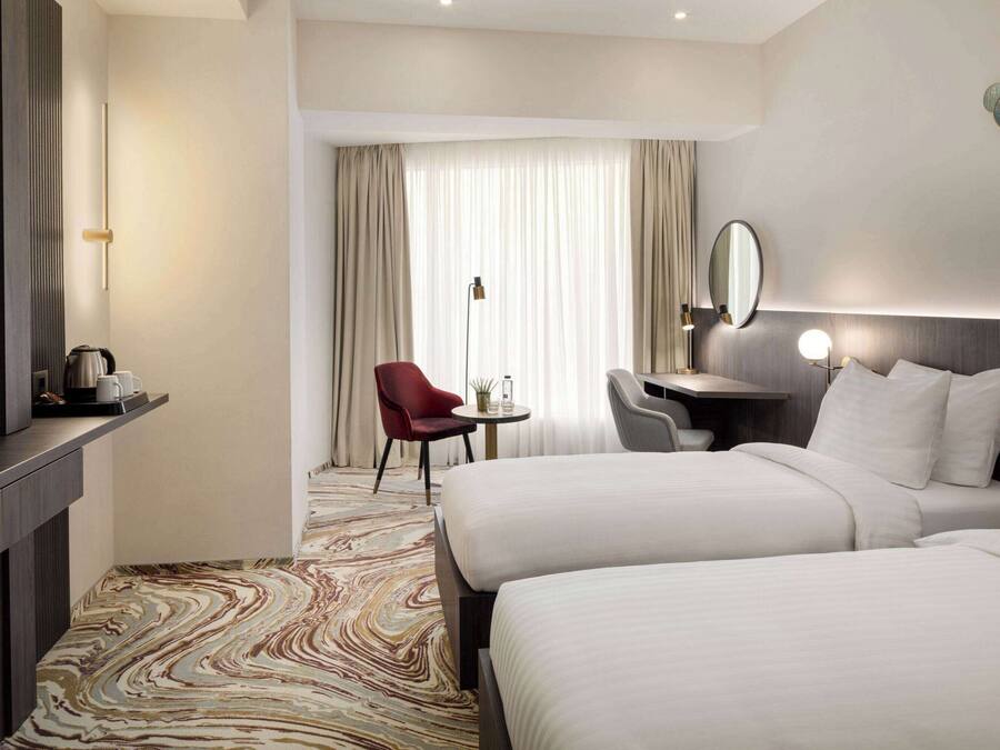 A hotel room with two beds, a desk, a chair, a TV, and a large window with curtains.