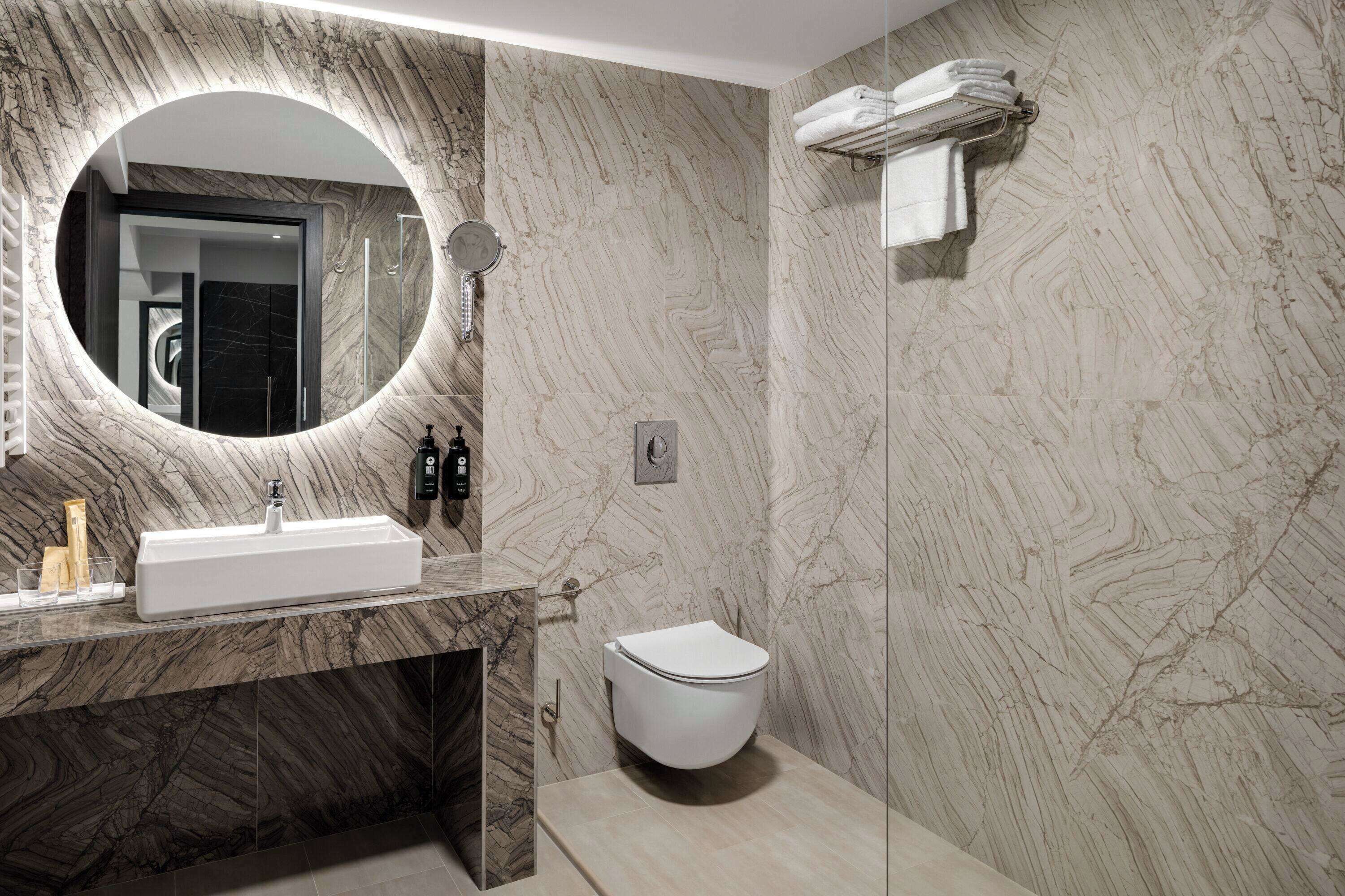 A modern bathroom with a marble vanity, a round mirror, a toilet, and a towel rack.