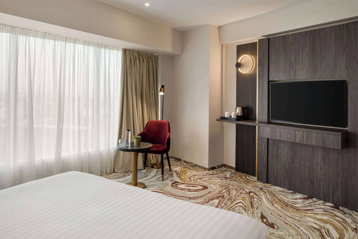 Habitación de hotel con una cama grande, una silla roja, una mesita con una lámpara, un televisor de pantalla plana montado en la pared y un piso con diseño de mármol.