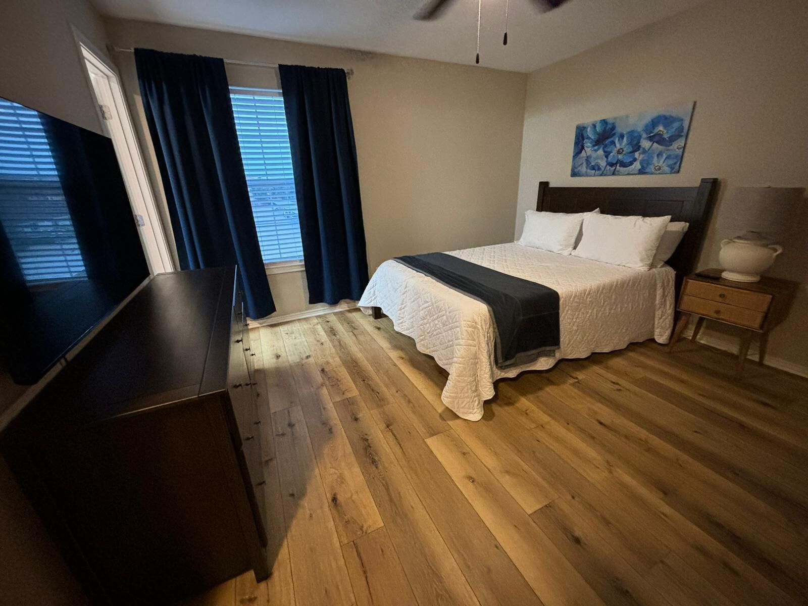 Une chambre à coucher avec un lit, une table de chevet, une lampe, un téléviseur et une fenêtre avec des rideaux.