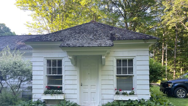 A small, single-story house with a white exterior and a dark roof. It features a front door, two windows with shutters, and a small porch area.