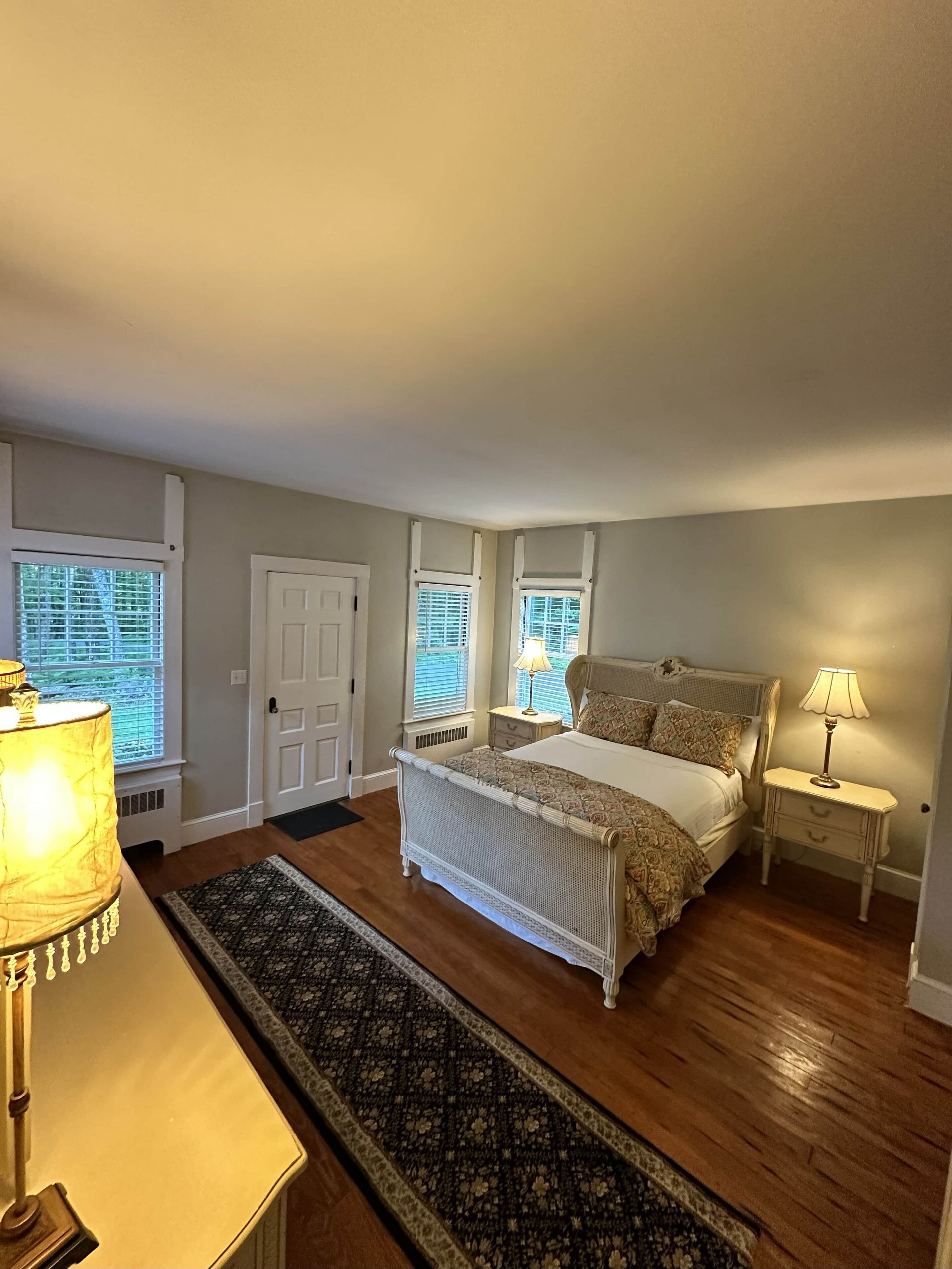 Une chambre à coucher avec un grand lit, deux tables de chevet avec des lampes, une fenêtre avec des stores et une porte.