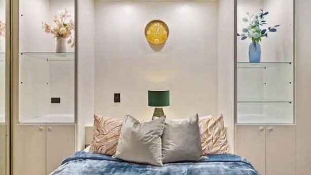 A bedroom with a bed, a wall clock, a lamp, and two glass shelves with vases.