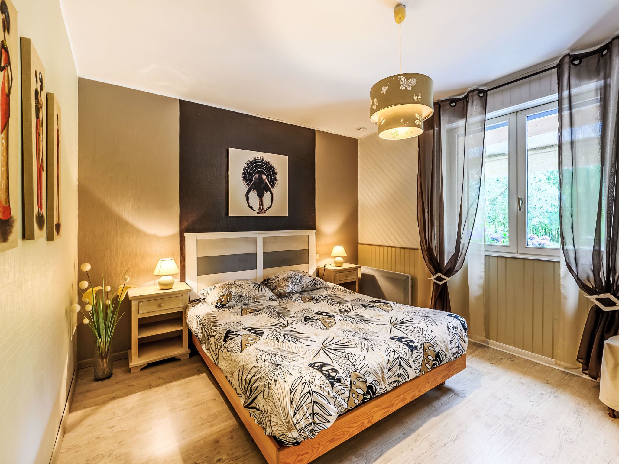 Une chambre à coucher comprenant un lit, des tables de chevet, une œuvre d’art accrochée au mur, une fenêtre avec des rideaux et un luminaire suspendu.