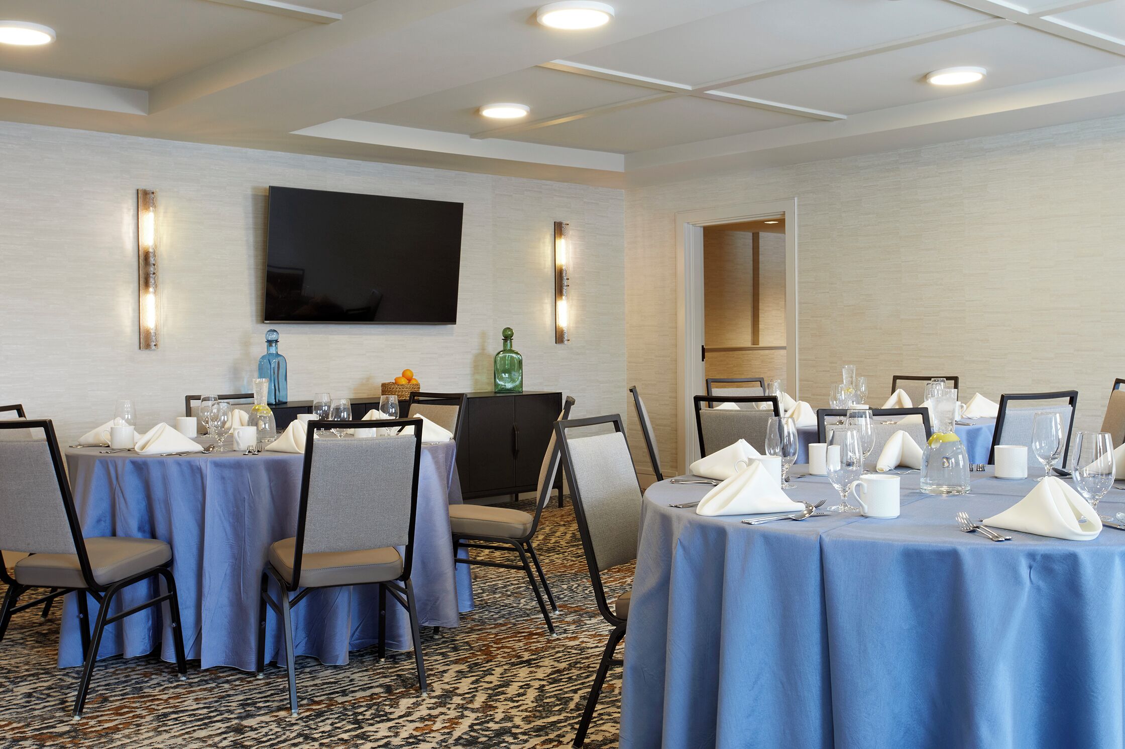 Un salón de conferencias con mesas redondas dispuestas para una reunión, equipado con sillas, mesas y diversos objetos como vasos y una botella.