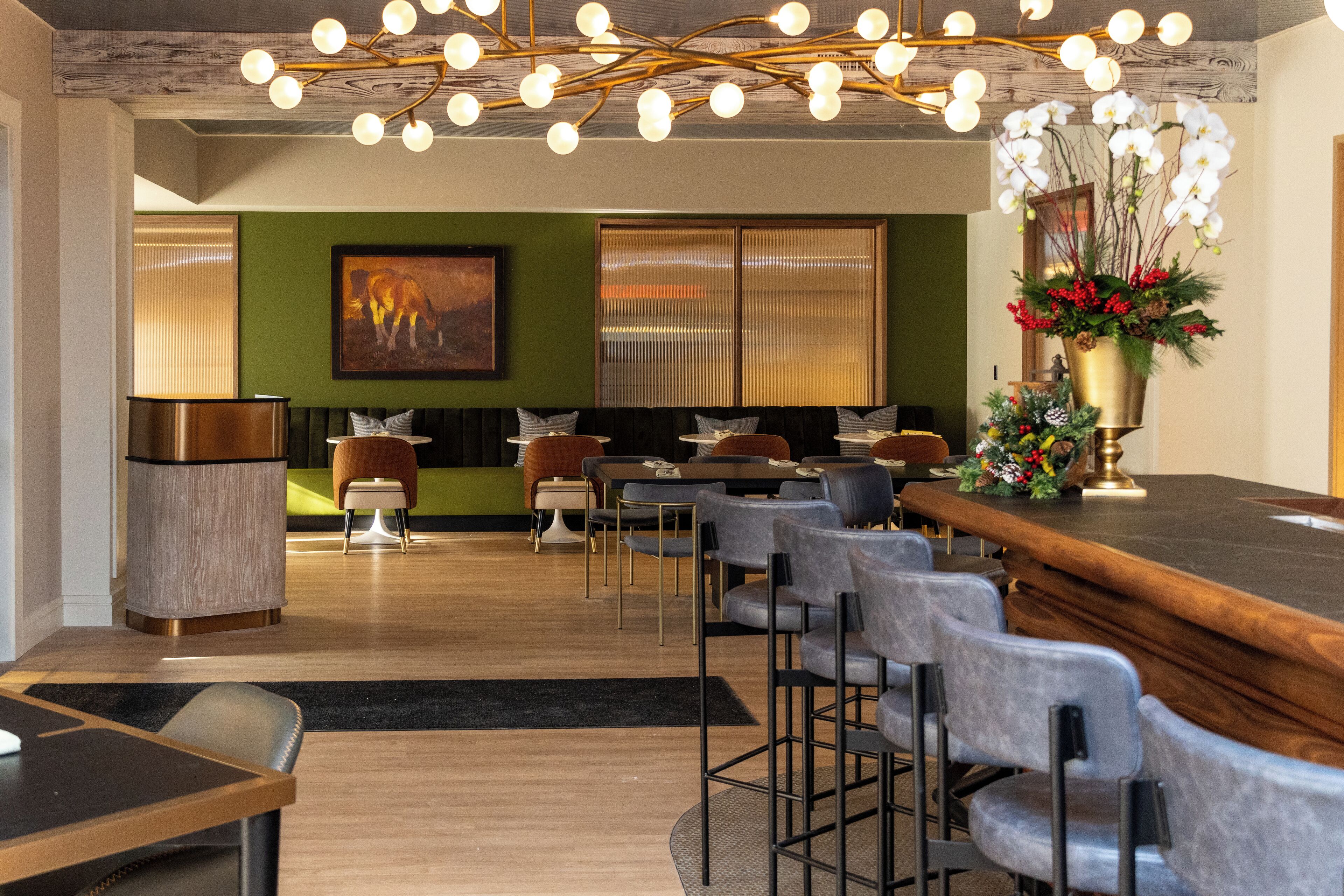 Un espace bar moderne avec un comptoir en bois et des tabourets de bar, un mur vert orné d’un tableau et une grande fenêtre avec des stores.