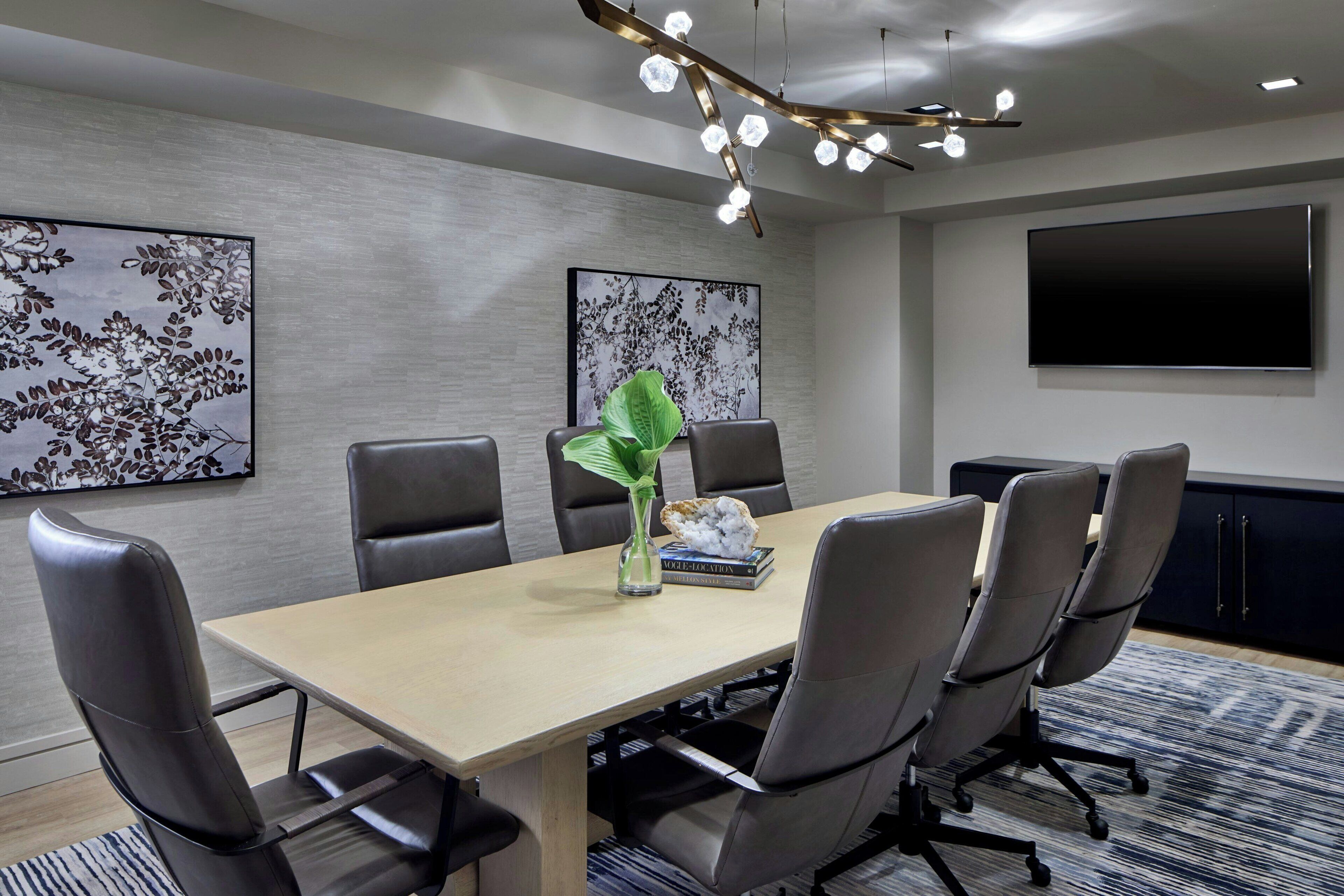 A conference room with a long table, six chairs, a large flat-screen TV, and two floral wall art pieces.