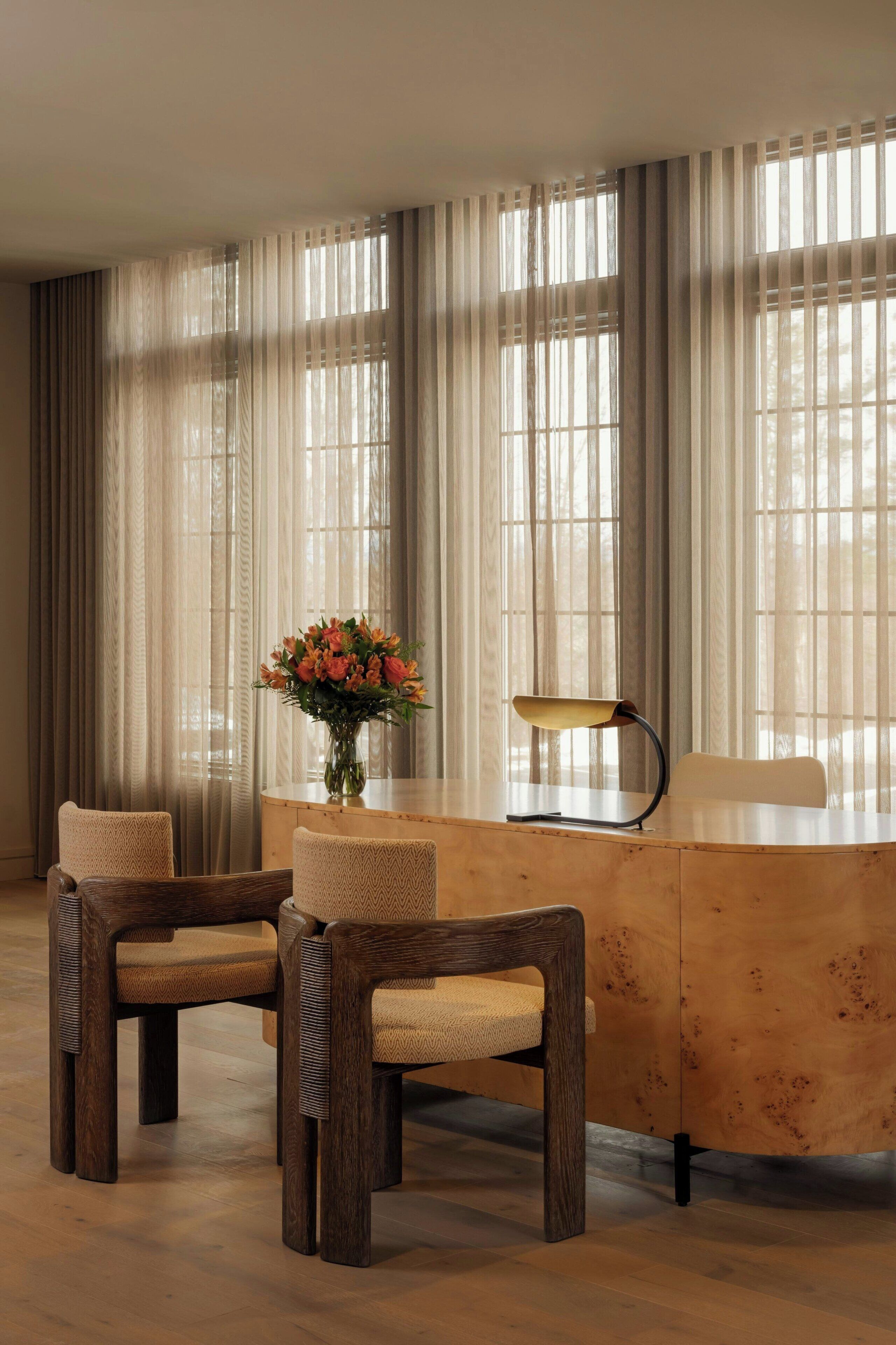 A room with a wooden dining table, two chairs, a vase with flowers, and sheer curtains.