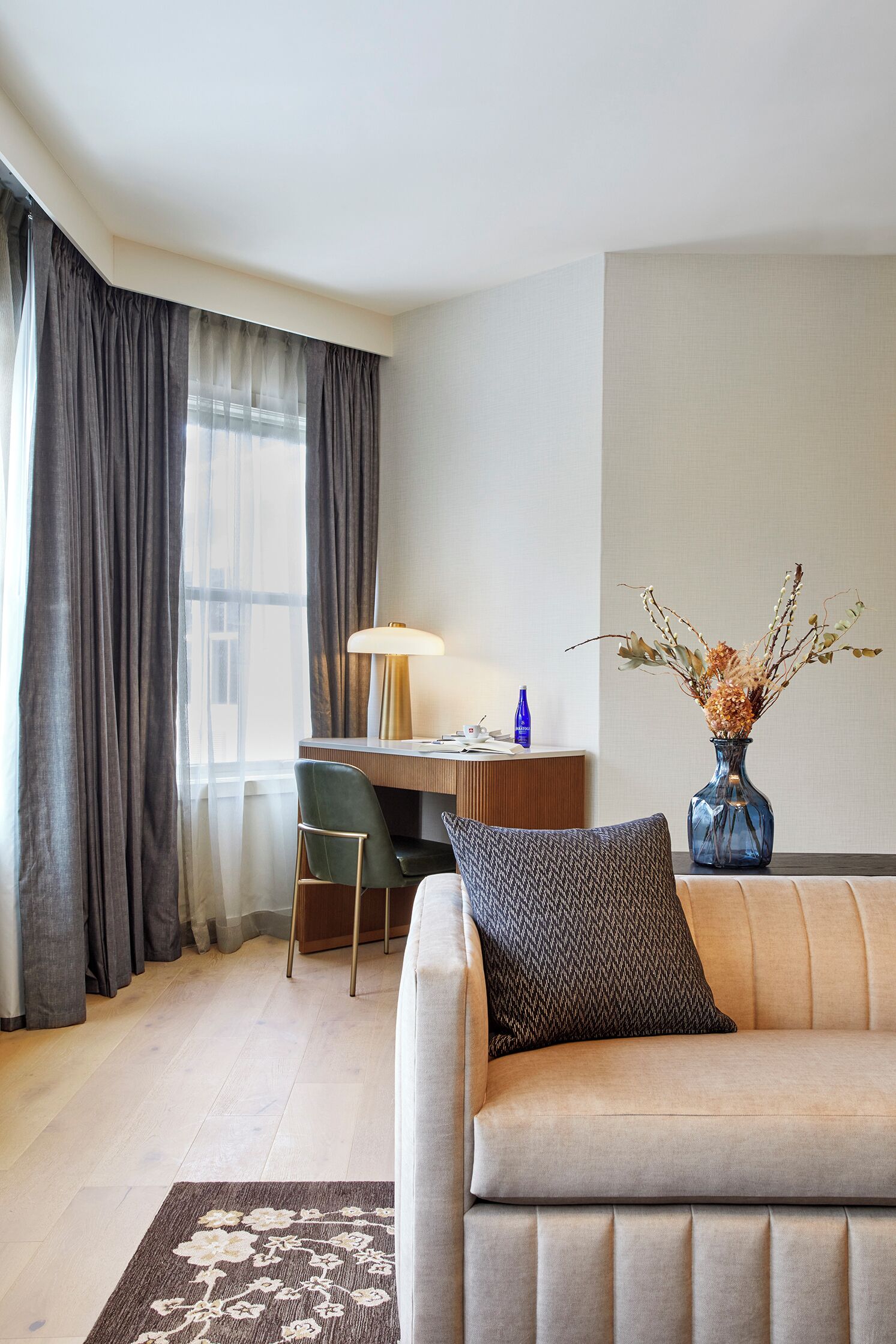 Hotellihuone, jossa on sohva, työpöytä, tuoli, lamppu ja maljakko kuivakukilla.