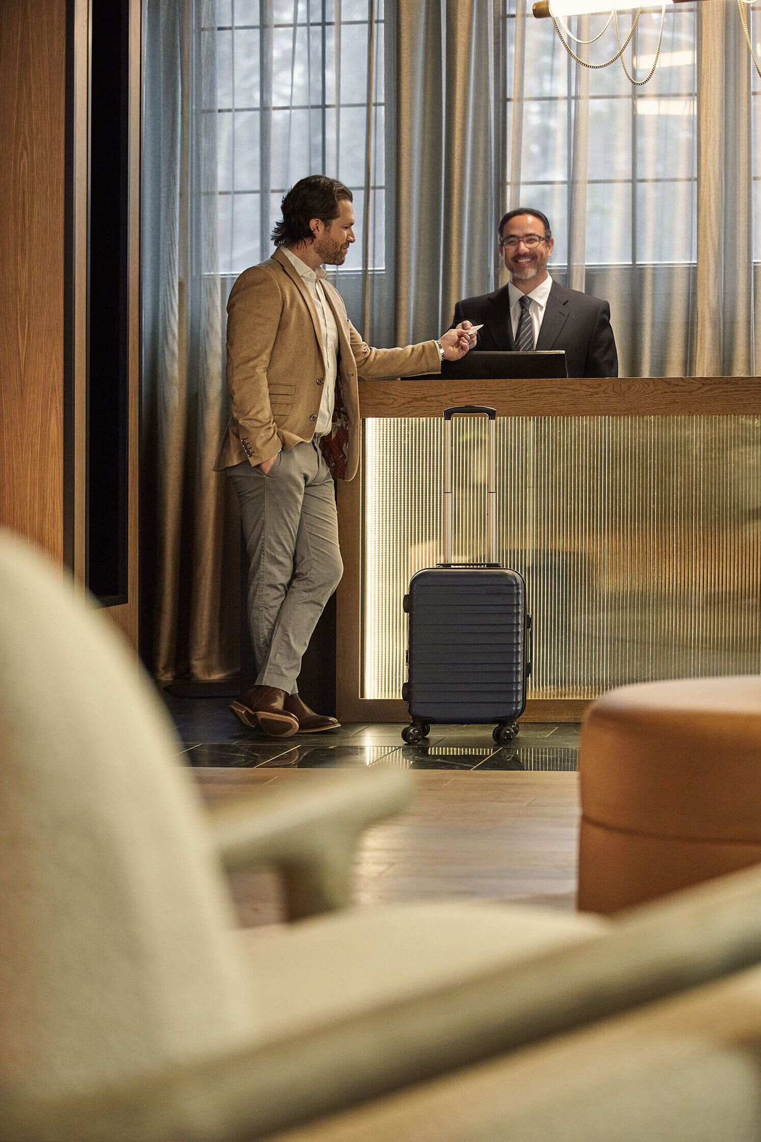 A man in a suit shaking hands with another man at a reception desk in a hotel lobby with large windows and a modern design.
