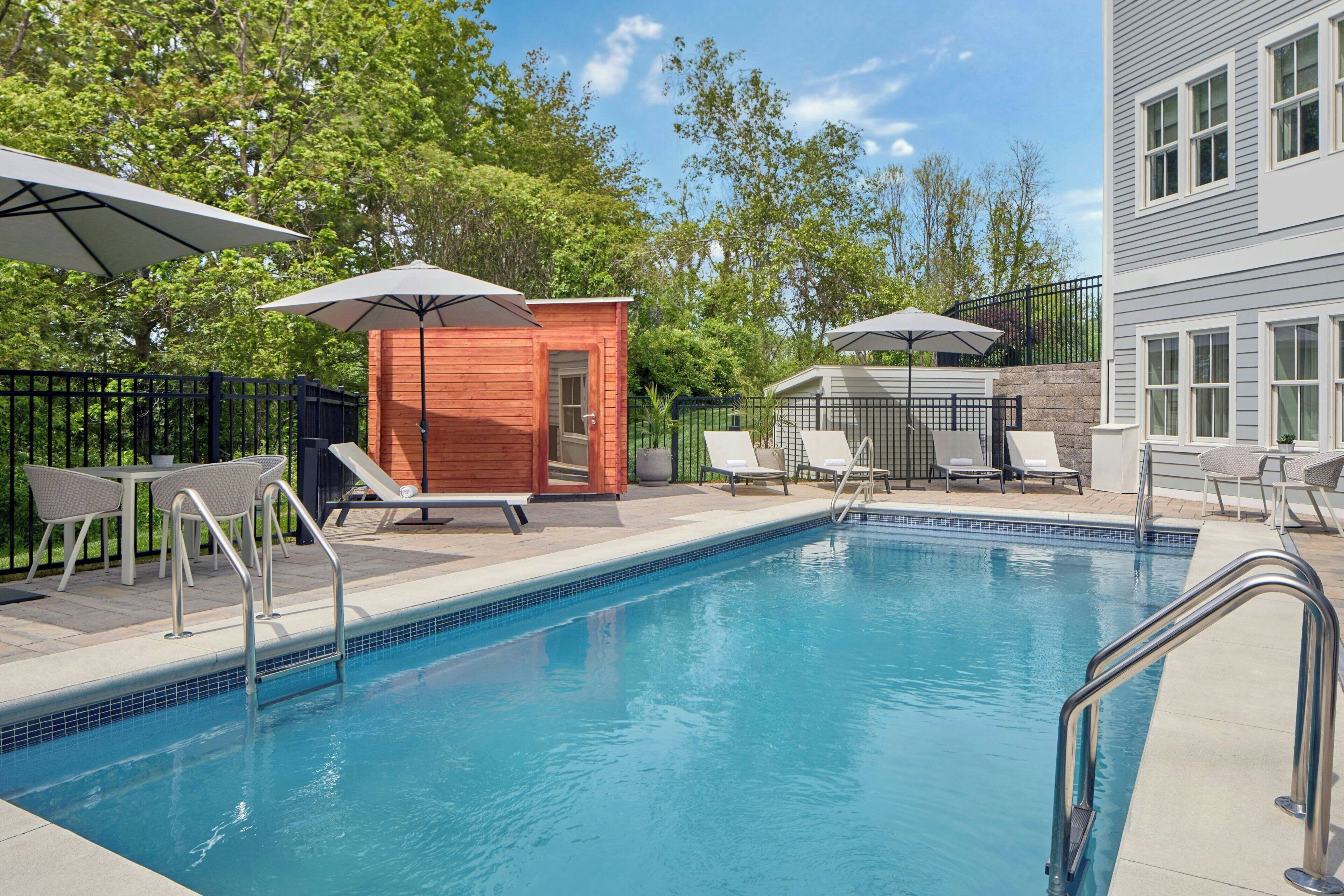A swimming pool with lounge chairs and umbrellas, surrounded by trees and a building.