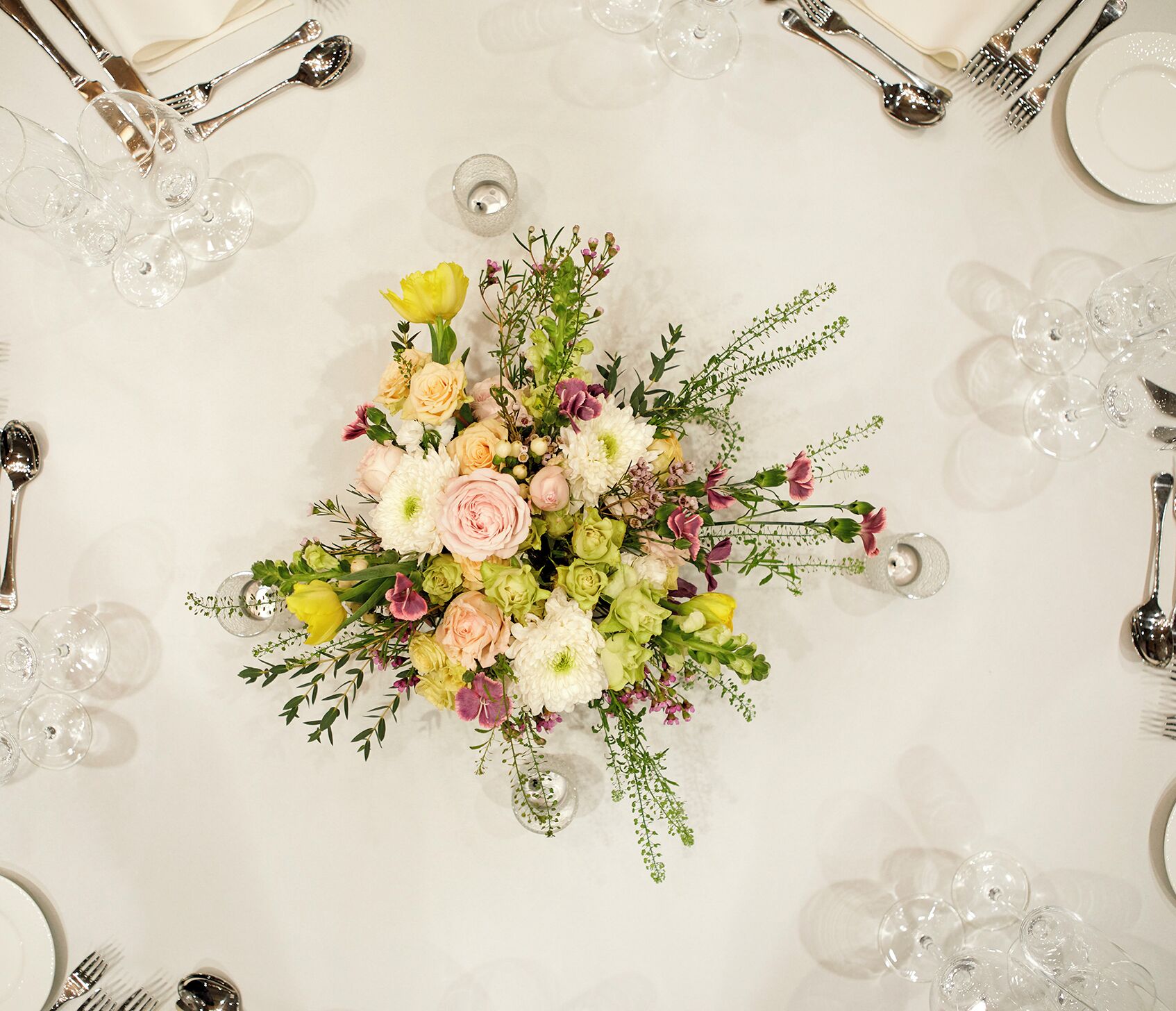 Et bord dækket med en blomsteropsætning, bestik og klare glas.