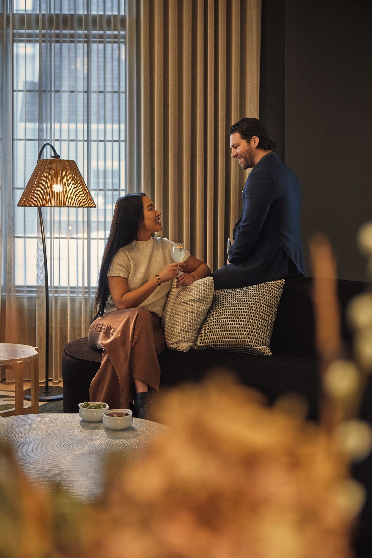 A couple sitting on a black sofa in a well-lit room with a large window, a floor lamp, and a small table with bowls.