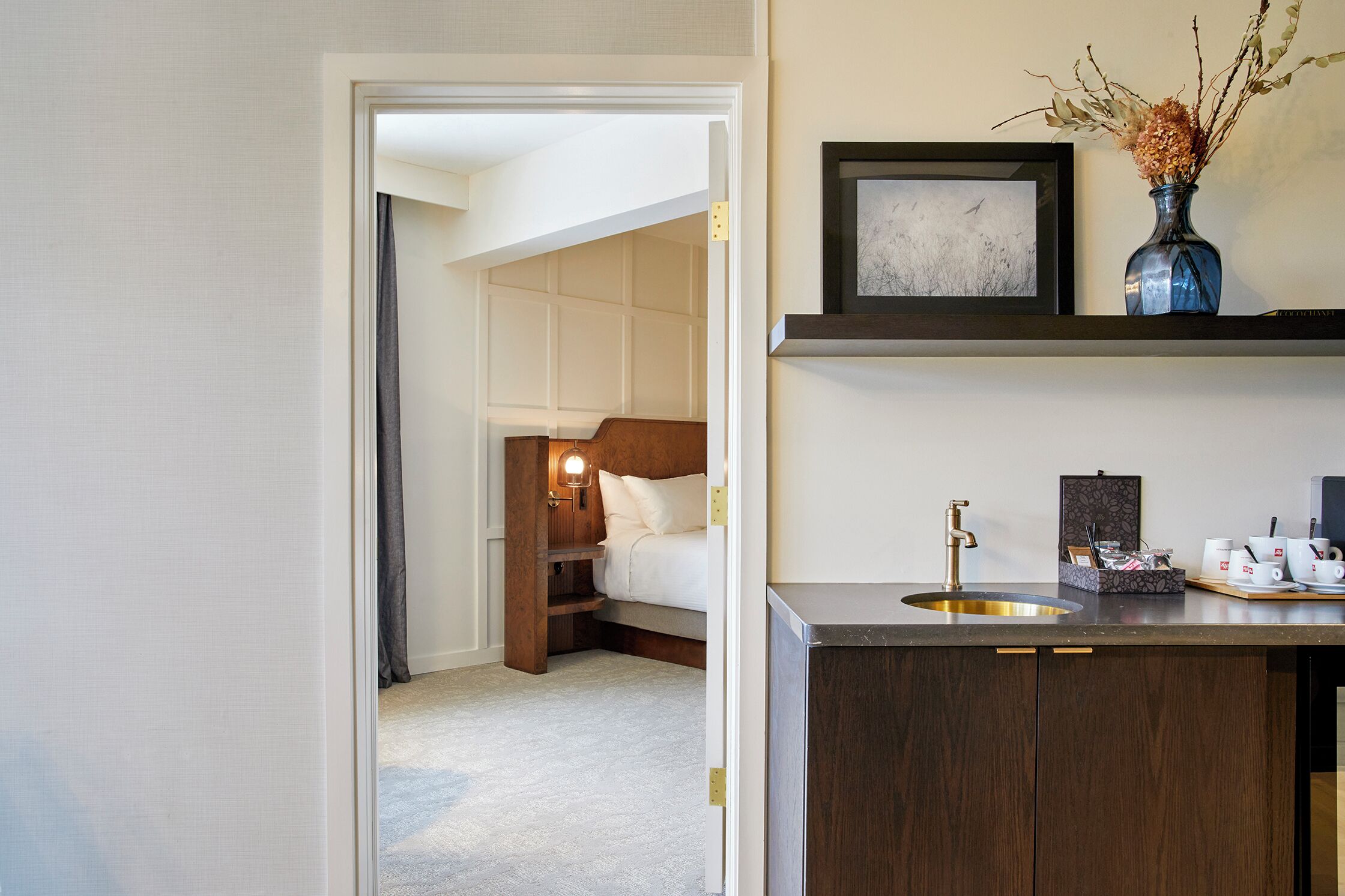 A hotel room with a bed, a bedside table, a nightstand, a built-in shelf, a sink, and a mirror.