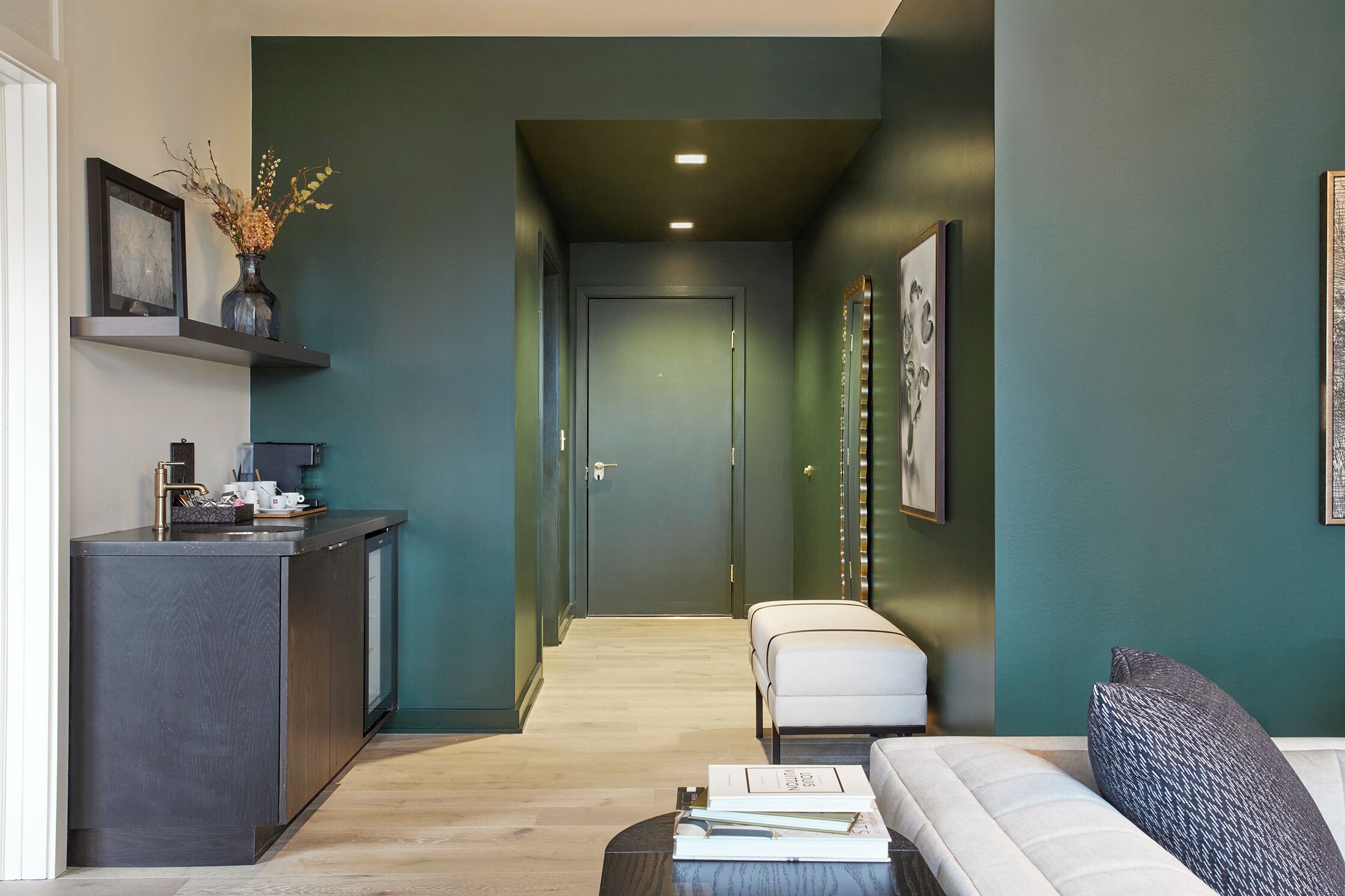 A modern interior with a dark green wall, a wooden floor, a white sofa, a small table with a vase, and a kitchen area with a dark countertop and a shelf with decorative items.