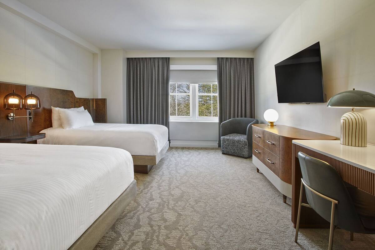 A hotel room with two beds, a desk, a chair, a TV, and a window with curtains.