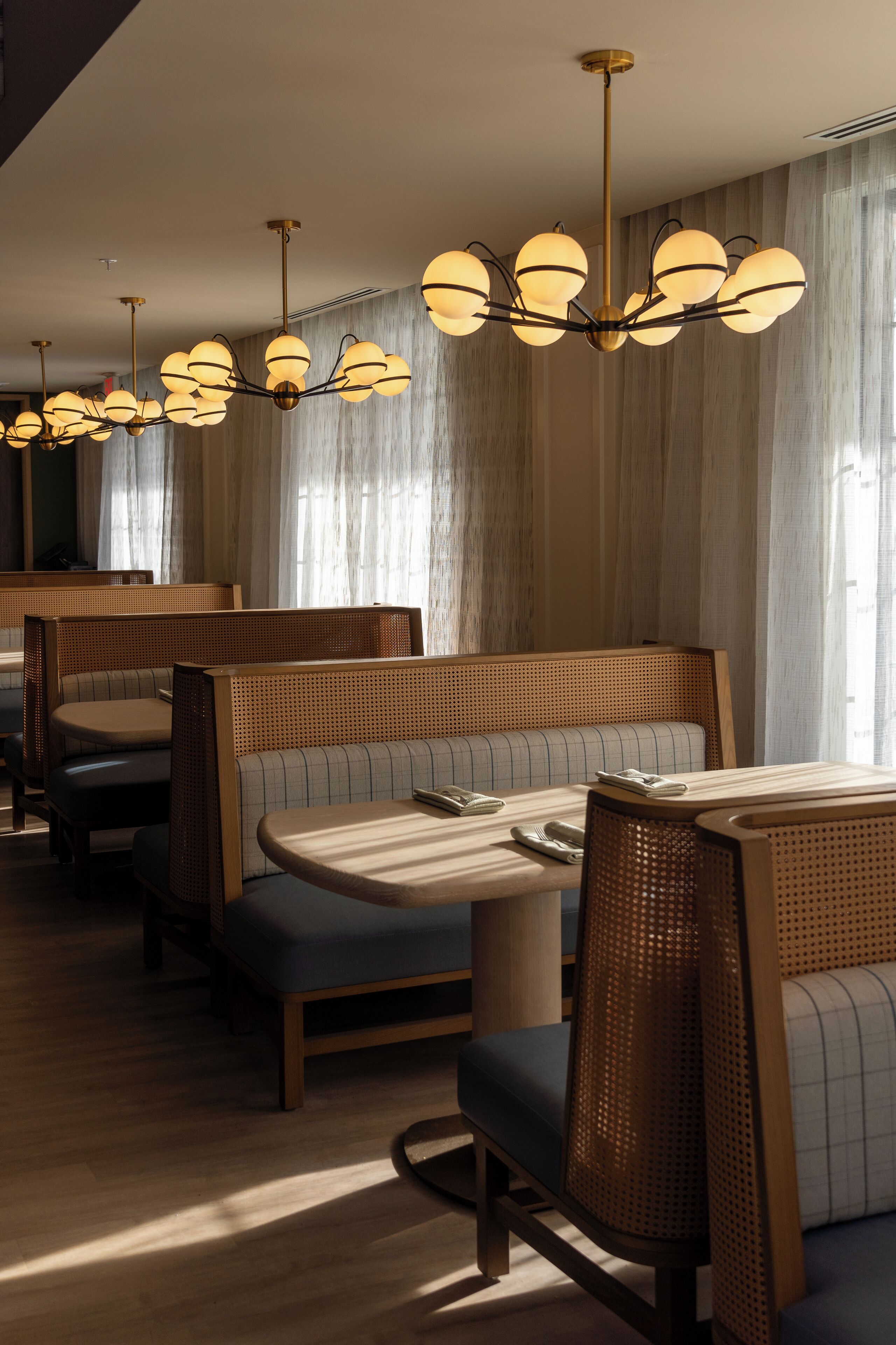 A hotel dining area with wooden booths and tables, a chandelier, and large windows with curtains.
