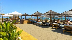 Een terras aan het strand met ligbedden en rieten parasols, met uitzicht op de zee.