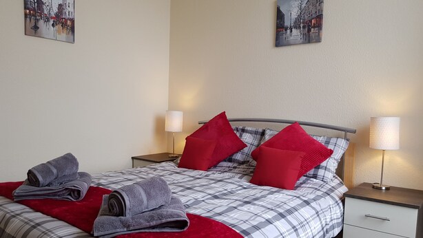 A neatly made bed with a plaid comforter, red pillows, and folded towels. There are two bedside tables with lamps, and artwork on the walls. - Charlotte Street (Ayr)