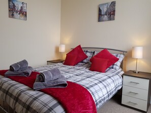 A neatly made bed with a plaid comforter, red pillows, and folded towels. There are two bedside tables with lamps, and artwork on the walls. - Charlotte Street (Ayr)