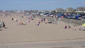 A crowded beach with people sunbathing and walking, and a parking area with cars. - Charlotte Street (Ayr)
