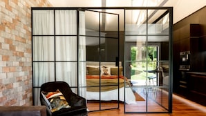 A modern bedroom with a large sliding glass door leading to a balcony, a bed with a decorative pillow, a black leather chair, and a wooden floor.