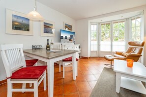 A dining area with a wooden table, white chairs with red cushions, a brown leather chair, and a small table with a glass. - Meeresleuchten - Ab 2025 bei uns im Angebot, für Max. 4 Personen (Sylt)