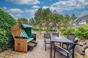 Eine Terrasse im Freien mit Korbstuhl, Tisch und Stühlen. Im Hintergrund sind Bäume und ein Auto zu sehen.