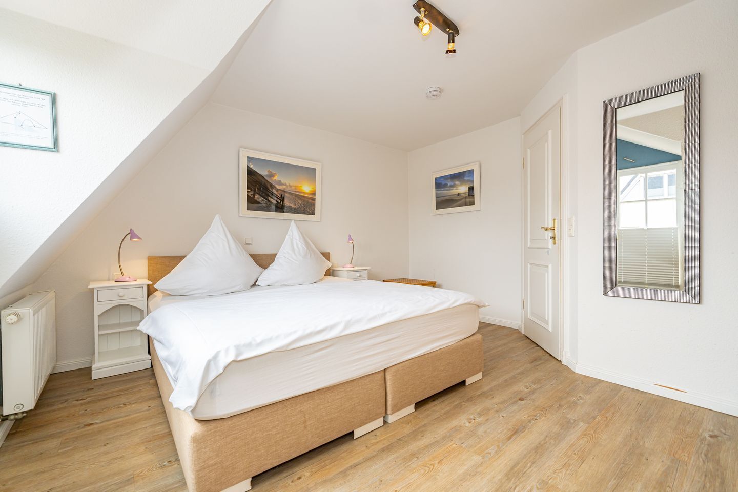 Une chambre à coucher avec un lit, deux tables de chevet, un grand miroir et deux tableaux encadrés accrochés au mur.